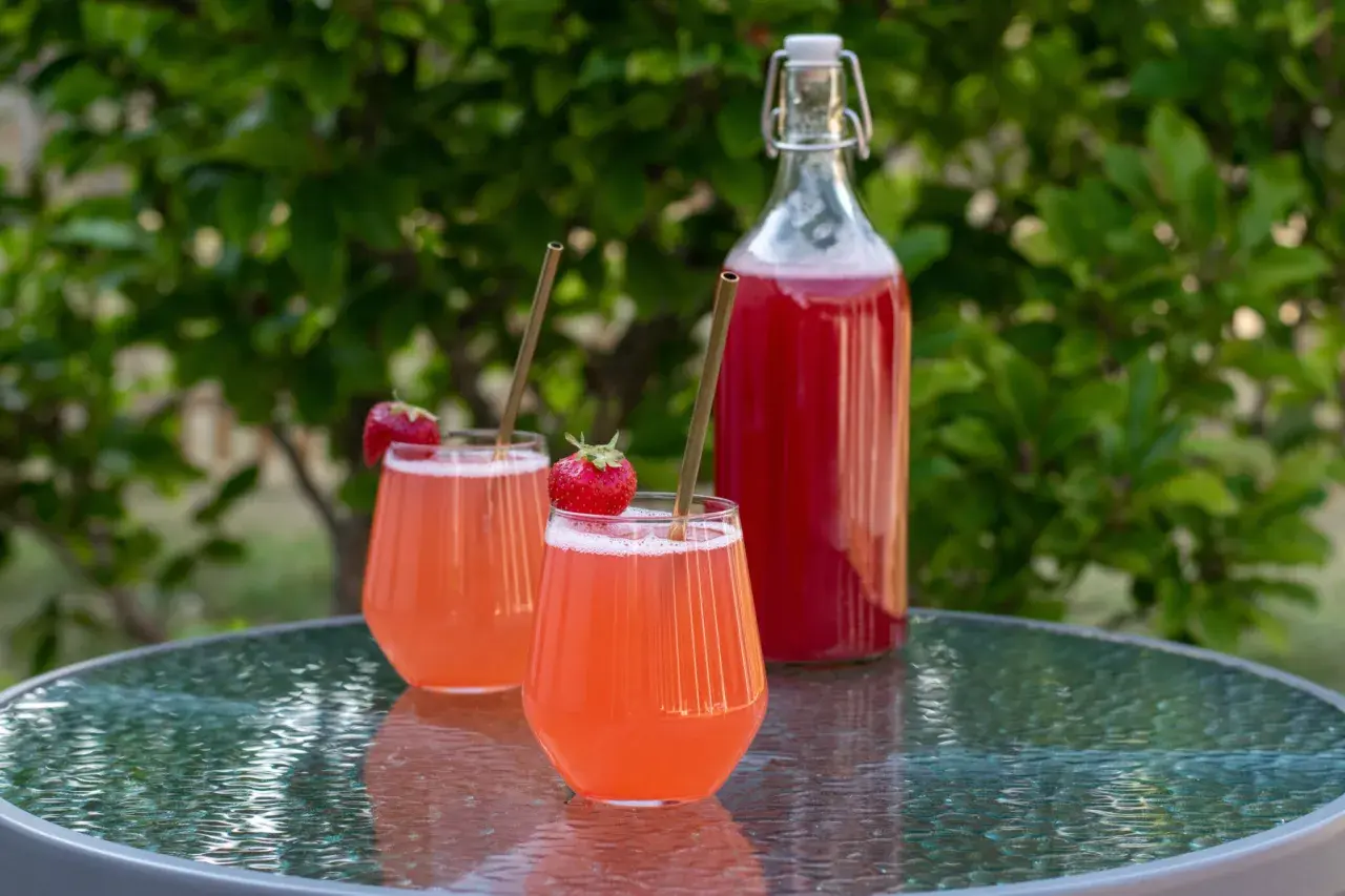 Två glas med rosa jordgubbssaft, garnerade med färska jordgubbar och sugrör, står på ett glasbord. En flaska med jordgubbssaft står bakom. Ett perfekt jordgubbssaft recept för sommaren.