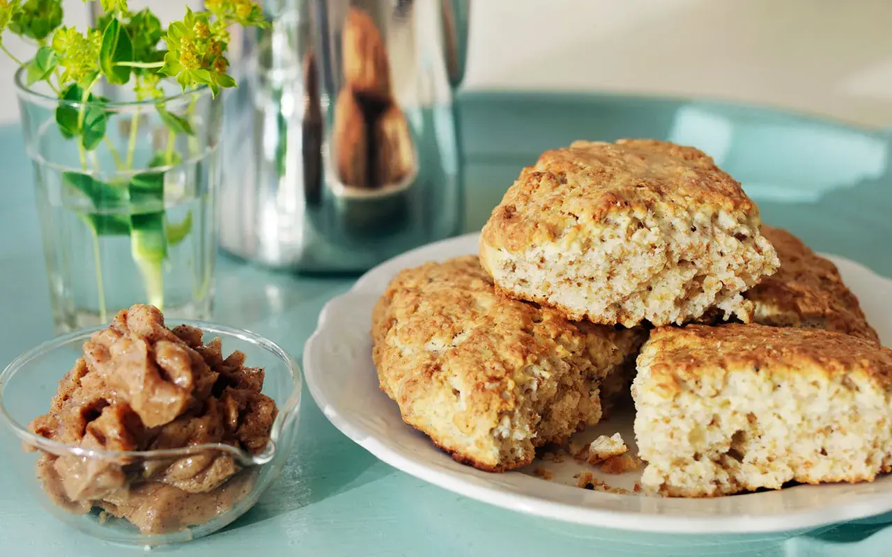 Nybakade kardemumma scones på en vit tallrik, med en skål fylld med kardemummafyllning och en vas med gröna blommor i bakgrunden.