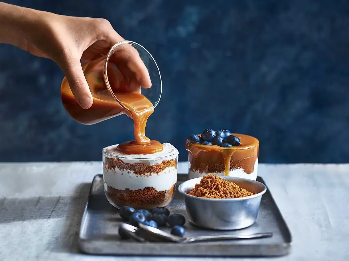 En hand häller karamellsås över en läcker cheesecake i glas. Blåbär och smulor fulländar desserten.