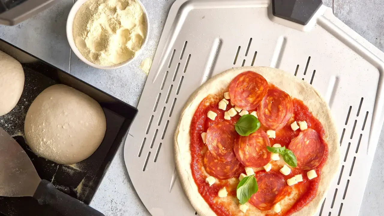 Pizzadeg recept: färdig pizzadeg med salami, ost och basilika på en pizzaspade. Bredvid ligger mer deg och en skål med mjöl.
