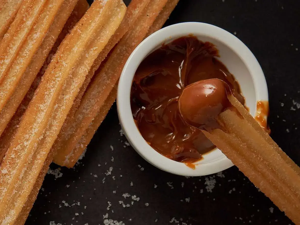 Churros coated in sugar being dipped into a bowl of dulce de leche.