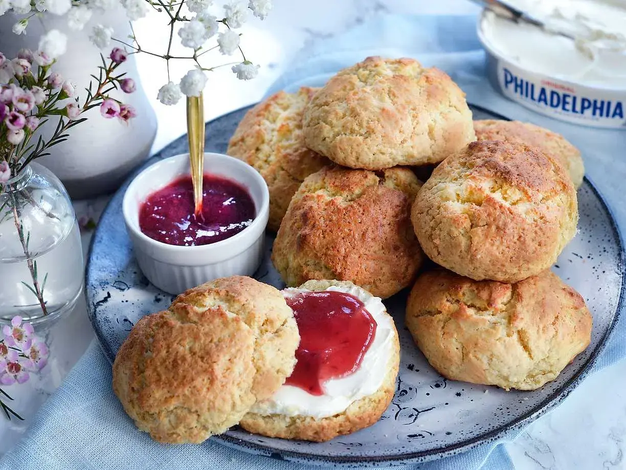 Färska scones bikarbonat på ett fat med sylt och grädde. En vacker bukett blommor i bakgrunden.