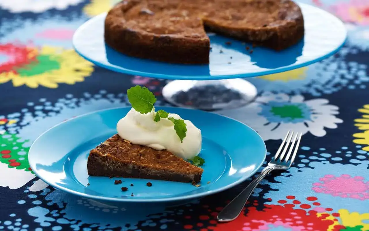 En bit kladdkaka med vispad grädde och mynta på en blå tallrik. Hela kladdkakan syns på en tårtfat i bakgrunden. Ett perfekt kladdkaka recept för fikat.