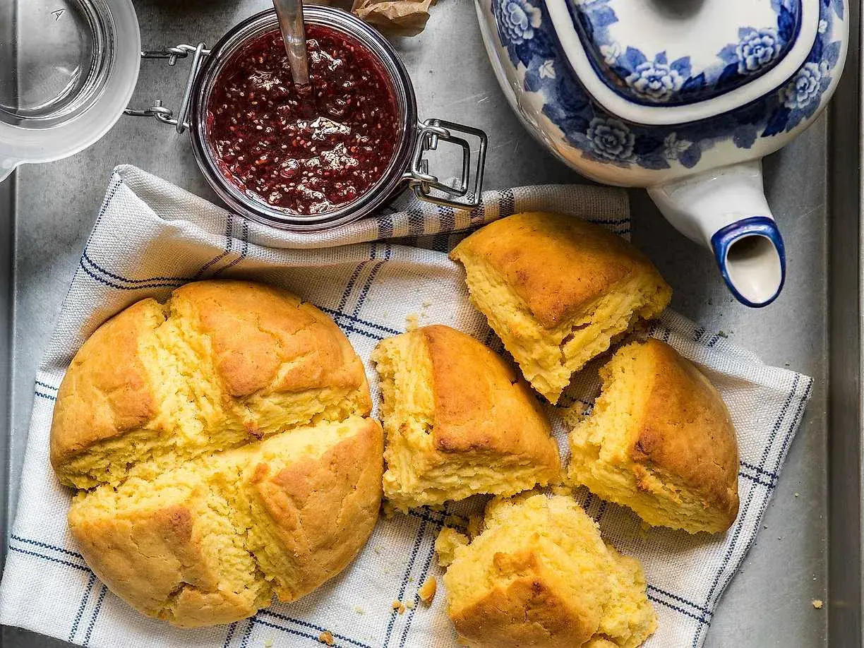 Gyllene scones majsmjöl, en burk hallonsylt och en blåvit tekanna. Perfekt för en mysig fika.