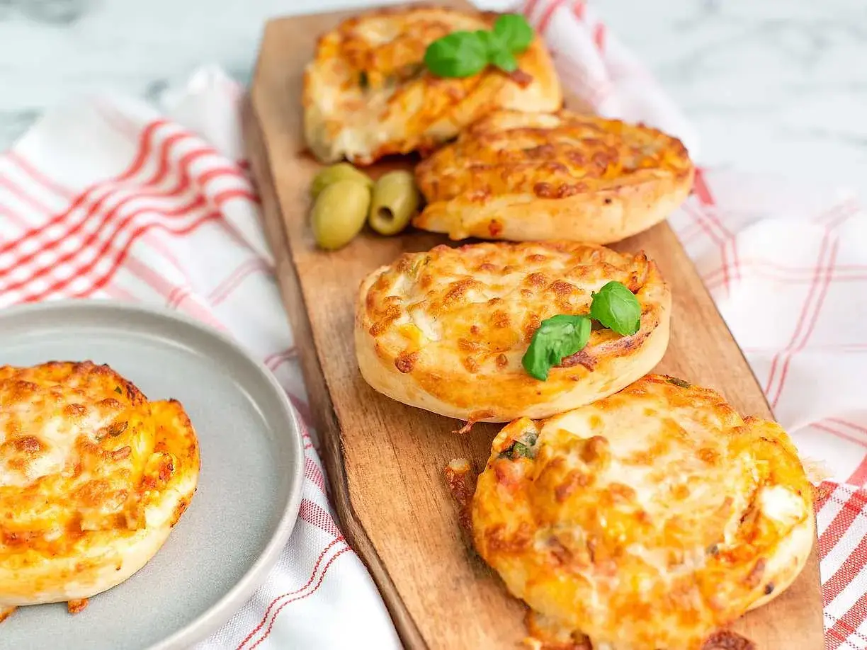 Four golden-brown pizza buns topped with melted cheese and basil, served on a wooden board with olives.