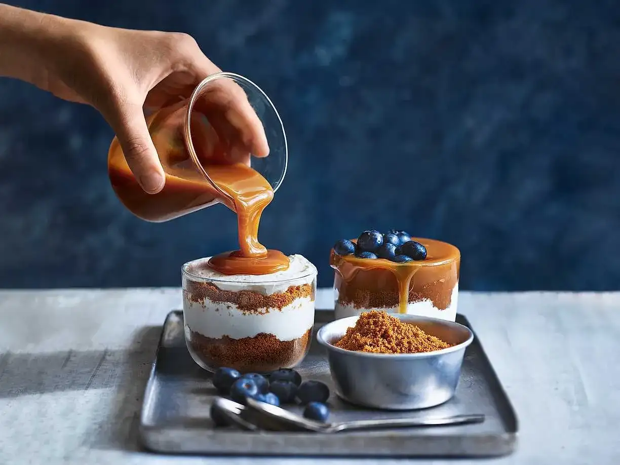 En hand häller karamellsås över en läcker cheesecake i glas. Blåbär och smulor fulländar desserten.