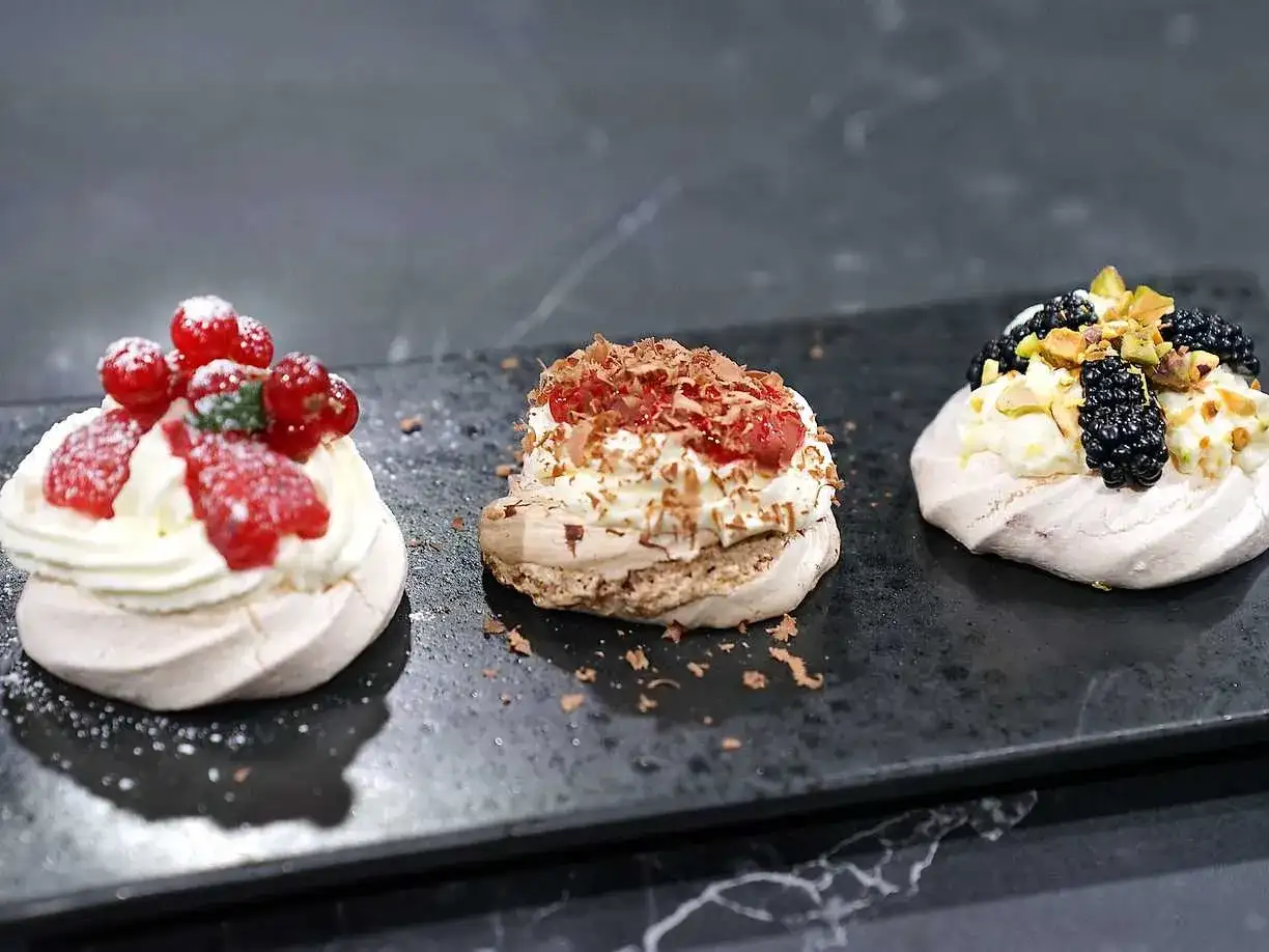 Tre små pavlovakakor med olika toppingar: jordgubbar och vinbär, choklad och hallon, samt björnbär och pistagenötter.