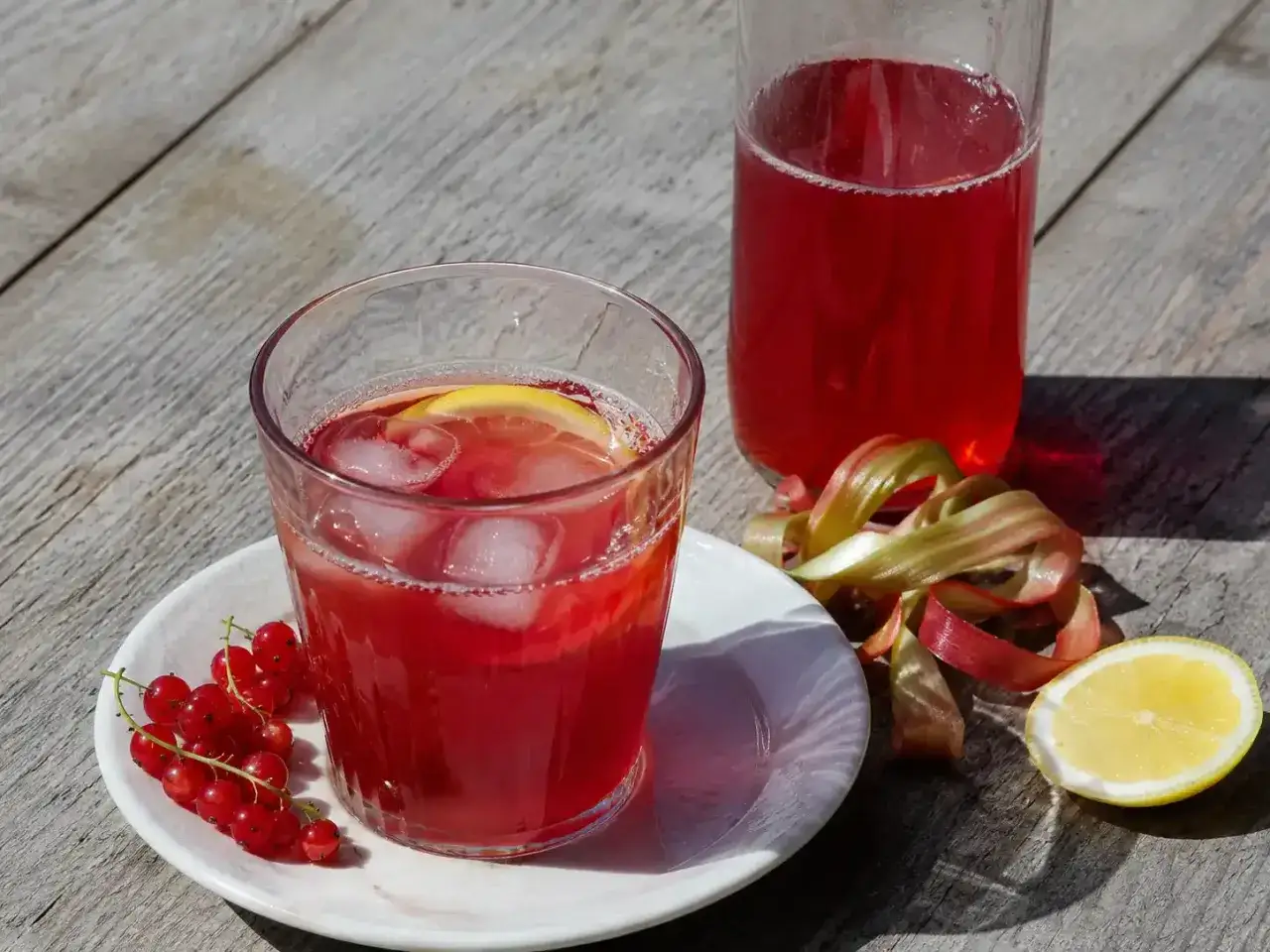 Ett glas med röd saft, is och citron, bredvid en flaska saft, vinbär och rabarber. Perfekt för att koka saft.