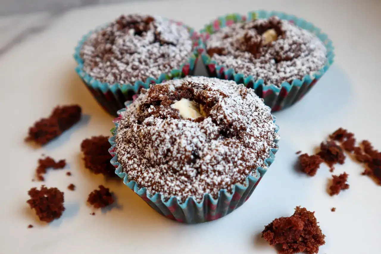 Tre glutenfria kladdkakemuffins med vit choklad i mitten, pudrade med florsocker. Några smulor ligger bredvid.