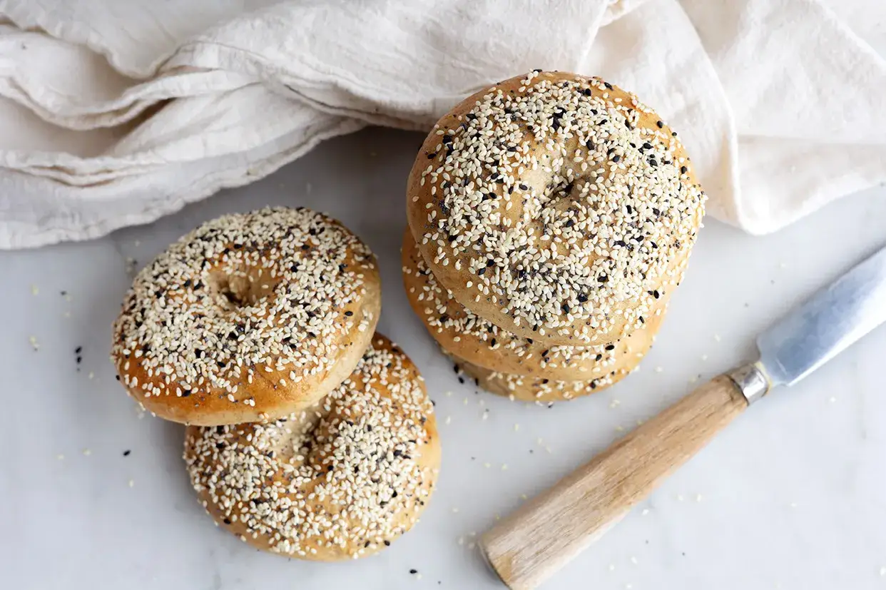 Färska bagels med sesamfrön och vallmofrön, redo för ditt bagels recept. En vit handduk och en kniv ligger bredvid.
