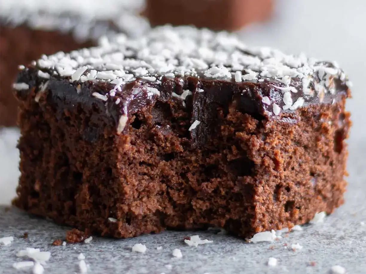 En saftig bit chokladkaka med glasyr och kokosflingor, som en del av en större sats mockarutor i långpanna.