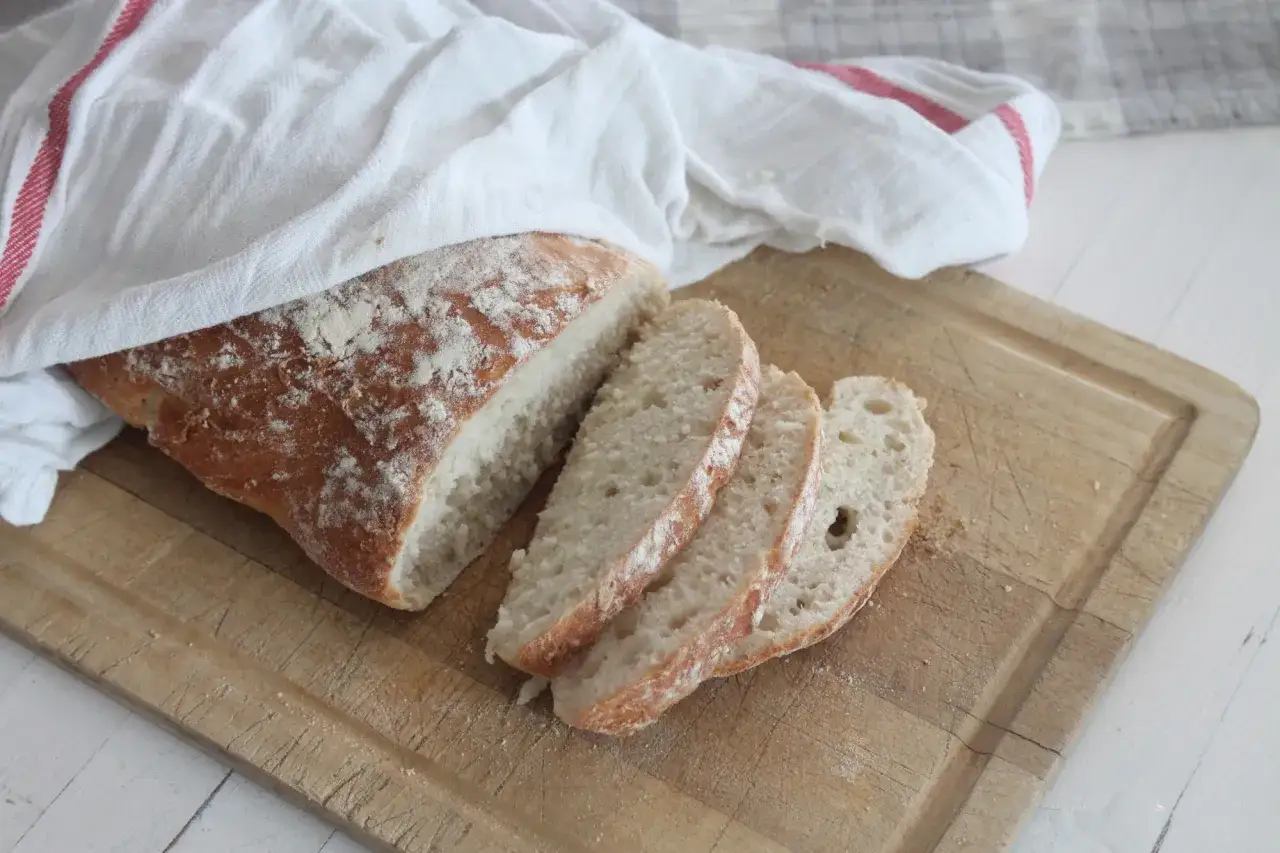 Nybakat br&ouml;d, t&auml;ckt med en vit handduk med r&ouml;da r&auml;nder, ligger p&aring; en sk&auml;rbr&auml;da. Br&ouml;det &auml;r skivat och visar en h&auml;rlig textur fr&aring;n kn&aring;dning.