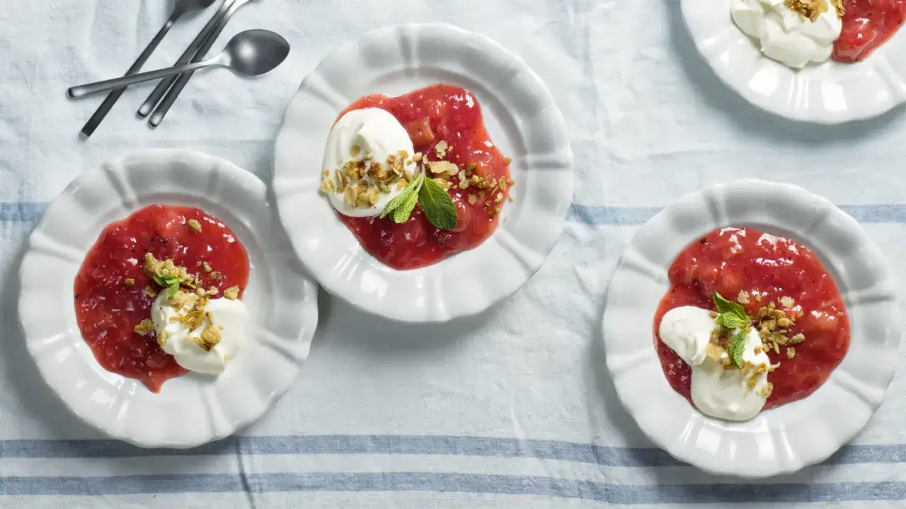 Tre vita skålar med rabarberkompott kardemumma, toppad med grädde och nötter. En härlig sommardessert.