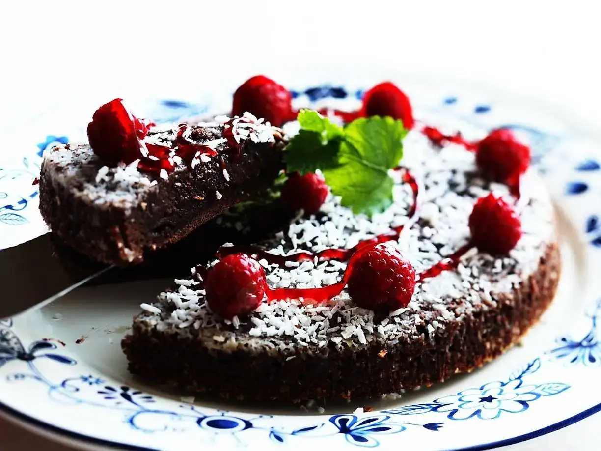 En stor kladdkaka med hallon, kokosflingor och röd sås på en blåvit tallrik. En bit är uppskuren.