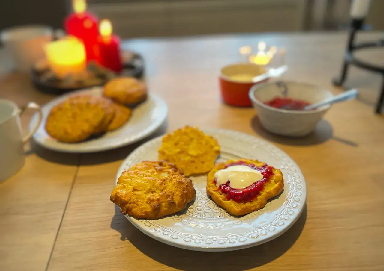 Gyllene scones med saffran, sylt och grädde på en tallrik. I bakgrunden syns röda ljus.