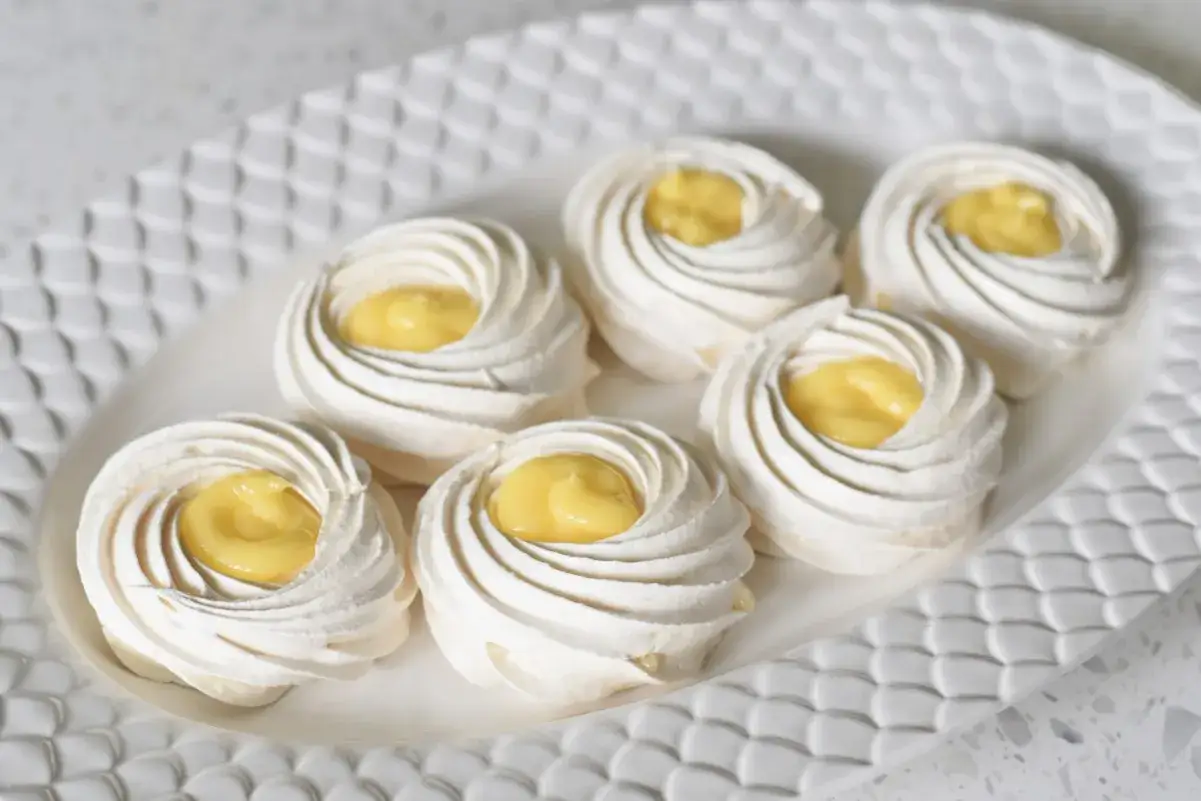 Sex små pavlova-bakelser med gul kräm i mitten, serverade på en vit, mönstrad tallrik.