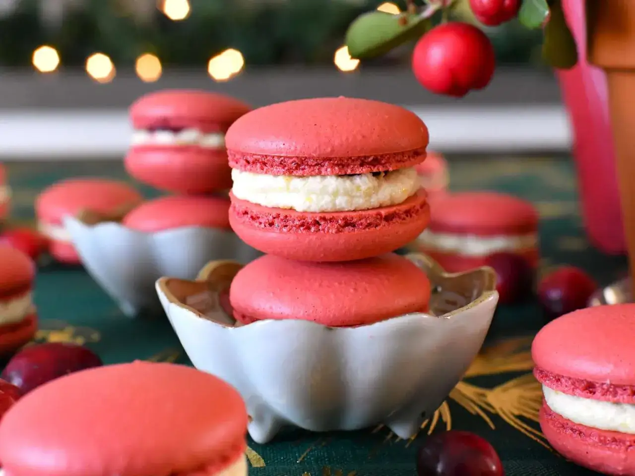 Rosa macarons med vit fyllning, perfekt f&ouml;r ett macarons recept. Dekorerade med r&ouml;da b&auml;r och julbelysning i bakgrunden.