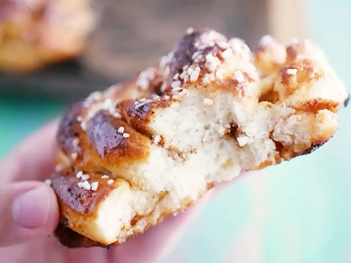 En hand h&aring;ller en bit av en gyllene, nybakad glutenfri kanelbulle, str&ouml;dd med p&auml;rlsocker.