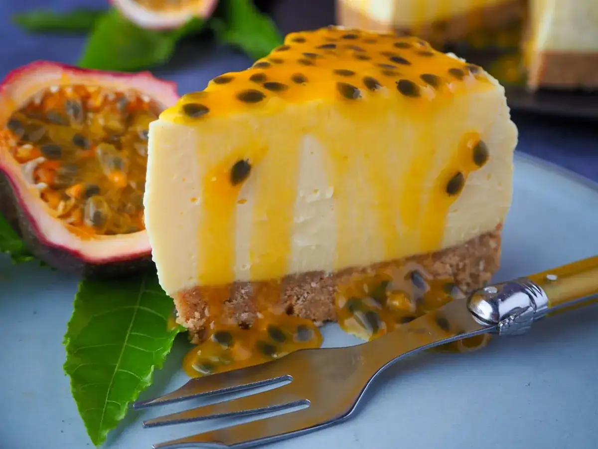 En krämig bit cheesecake med passionsfrukt, toppad med en glansig passionsfruktsås och frön. En halv passionsfrukt ligger bredvid.
