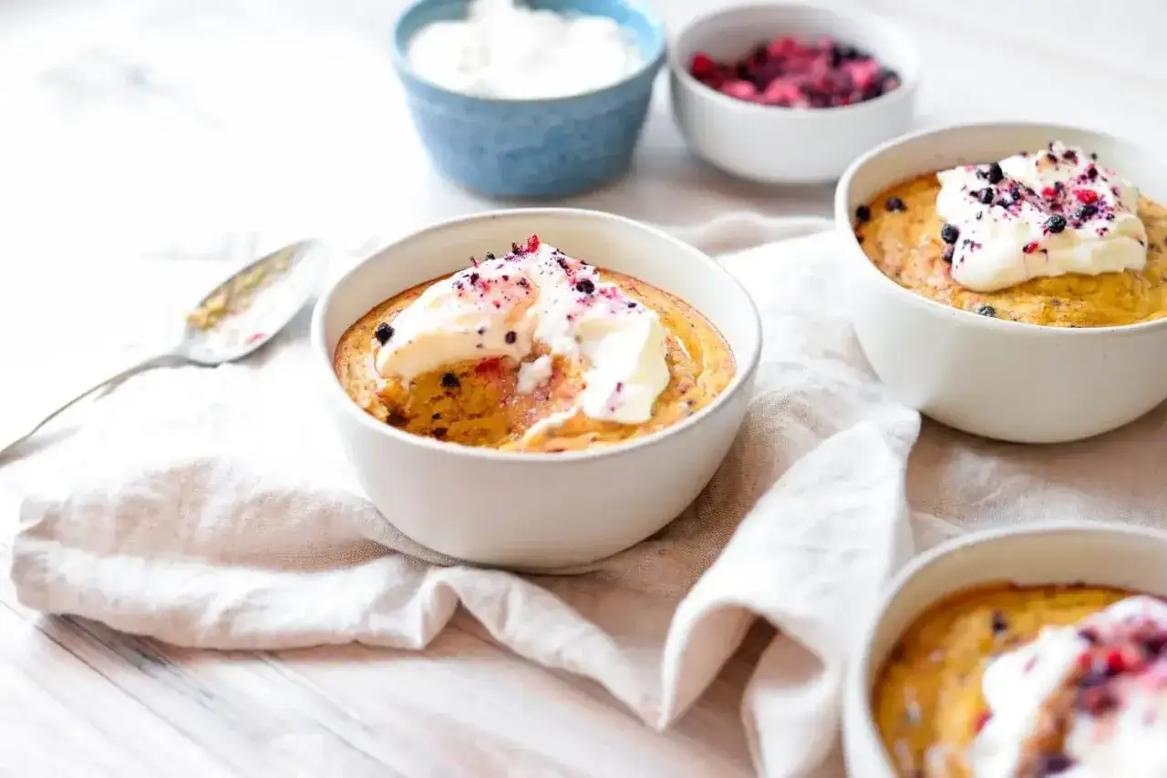 Tre skålar med fluffiga baked oats, toppade med grädde och bär. En sked ligger bredvid, redo för en god frukost.