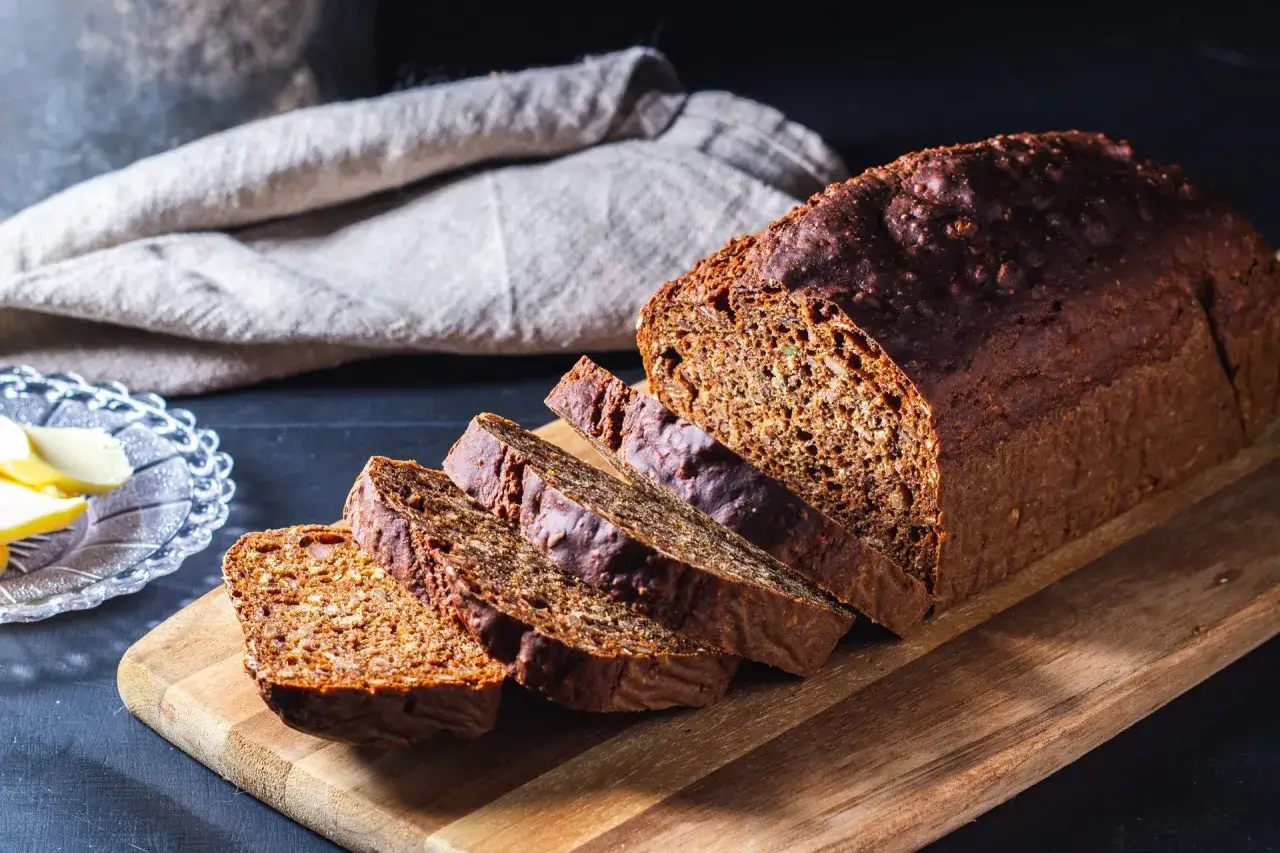 Mörkt, saftigt rågbröd skuret i skivor, perfekt för att baka med bikarbonat. Smör på en vacker skål i bakgrunden.