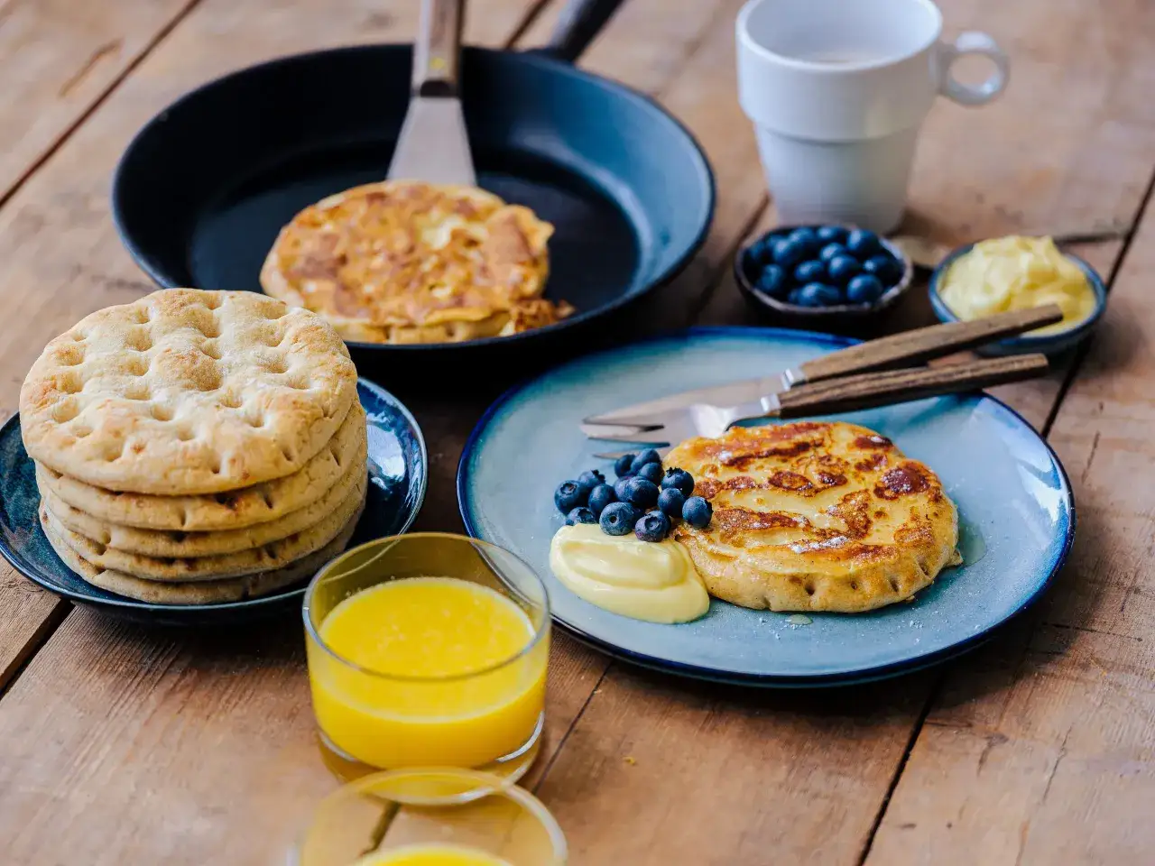 Fattiga riddare med blåbär och vaniljkräm på en blå tallrik, bredvid en hög med tunna pannkakor och ett glas apelsinjuice.