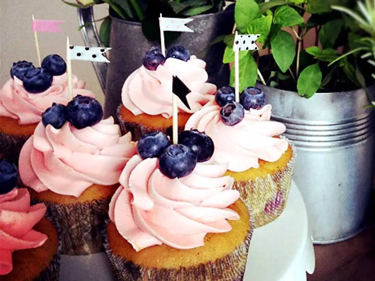 Sju muffins med rosa frosting och blåbär, dekorerade med små flaggor. Perfekt för ett sommarfika!