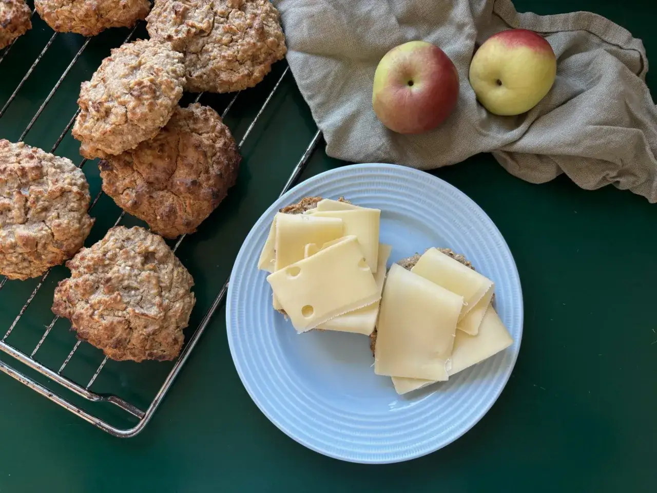 Nybakade &auml;ppelscones p&aring; ett galler, bredvid tv&aring; &auml;pplen och en sm&ouml;rg&aring;s med ost.