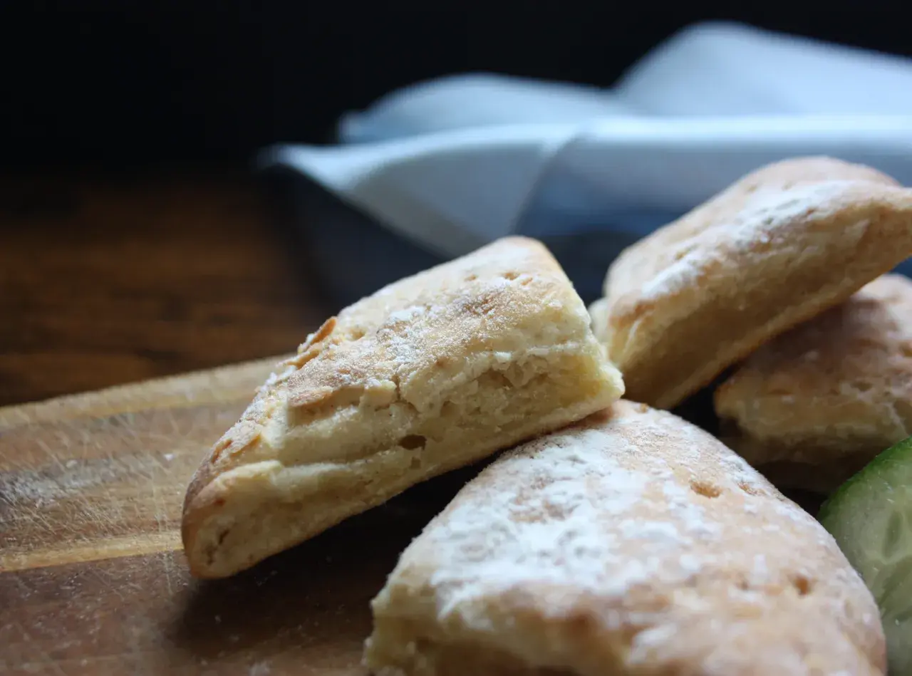Nybakade, enkla scones med pudrat socker på en träskärbräda. En bit gurka syns i bakgrunden.
