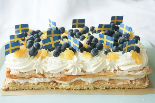 En stor brittatårta i långpanna, dekorerad med grädde, blåbär, apelsinbitar och små svenska flaggor. Perfekt för nationaldagsfirandet!