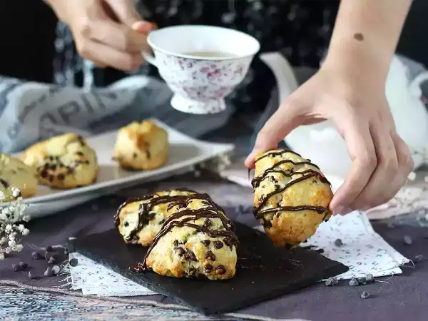 En hand plockar upp en chokladscone, dekorerad med chokladglasyr. Fler chokladscones ligger på ett fat.