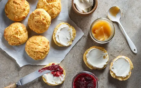 Nybakade glutenfria scones ligger på bakplåtspapper. Några är toppade med grädde och sylt, andra väntar på pålägg. Bredvid finns burkar med sylt och honung.