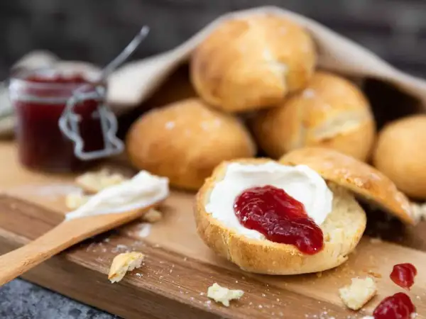 En sked med grädde och sylt på en halverad scones, redo att avnjutas. En burk sylt och fler av dessa bästa scones i bakgrunden.