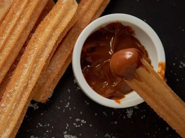 Churros coated in sugar being dipped into a bowl of dulce de leche.