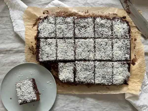 En plåt med chokladkakor, toppade med kokosflingor, redo att avnjutas som kärleksmums på påsk. En bit ligger på en tallrik.