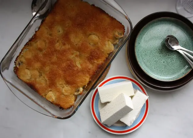 En gyllene bananpaj i en glasform, bredvid en hög med tallrikar och en liten tallrik med bitar av vit choklad.